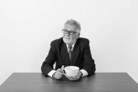 Studio shot of handsome senior bearded businessman thinking and holding coffee cup while sitting on wooden table against white backgroundの写真素材