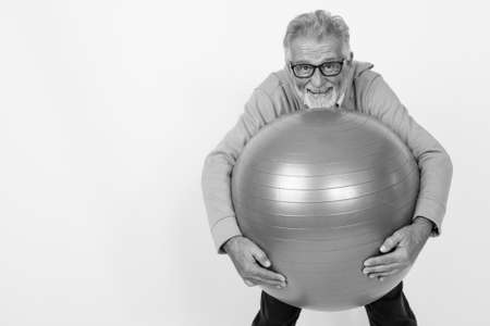 Studio shot of happy senior bearded man smiling while standing and holding gym ball ready for gym against white backgroundの写真素材