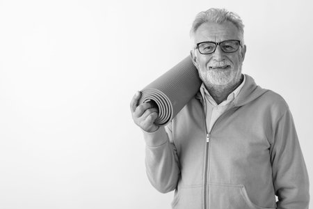 Studio shot of happy senior bearded man smiling while holding yoga mat ready for gym against white backgroundの写真素材