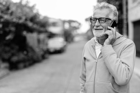 Happy handsome senior bearded man smiling while talking on mobile phone and thinking with eyeglasses...