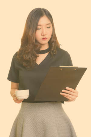 Young beautiful Asian businesswoman reading clipboard while holding coffee cupの写真素材