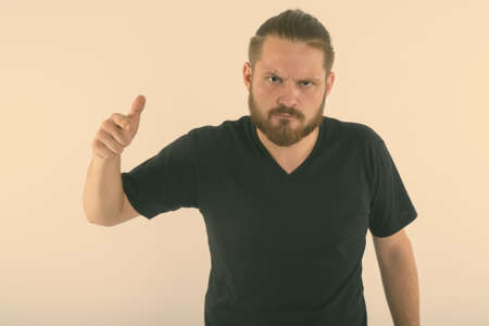Studio shot of young angry bearded man pointing at camera with hair tied against white backgroundの写真素材