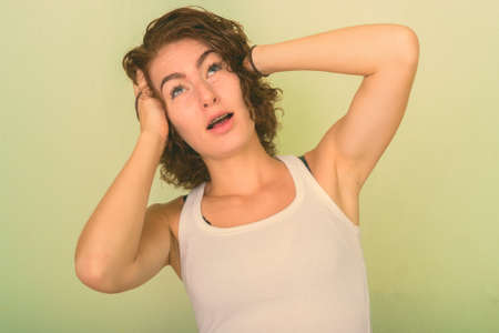 Studio shot of beautiful teenage girl thinking with both hands on head against green painted wallの写真素材