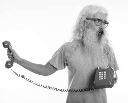 Studio shot of senior bearded man holding old telephone while looking away and annoyedの写真素材