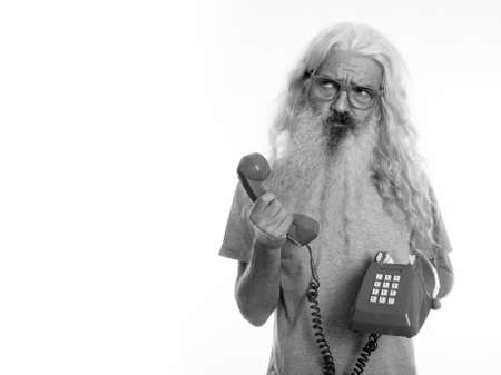 Studio shot of senior bearded man holding old telephone while thinking and looking upの写真素材