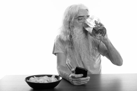 Studio shot of senior bearded man using mobile phone while drinking glass of beer with bowl of...