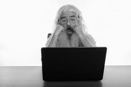 Studio shot of senior bearded man looking shocked while using laptop on wooden table