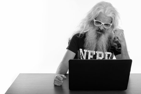 Studio shot of senior bearded nerd man looking angry while using laptop on wooden table