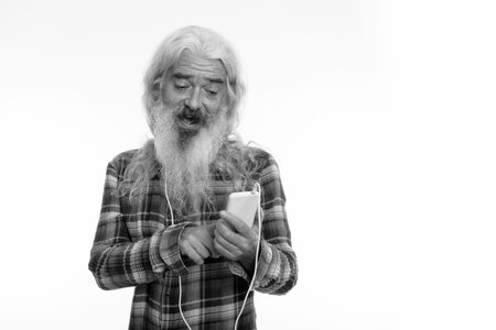 Studio shot of happy senior bearded man smiling while wearing headphones around neck and using...