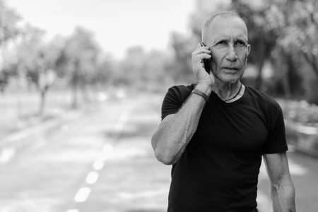 Bald senior tourist man thinking while talking on mobile phone on the side of the street at peaceful park in Bangkok Thailandの写真素材