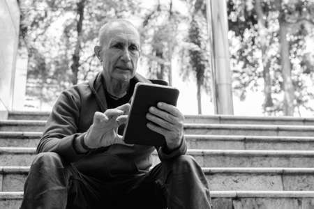 Bald senior man using digital tablet while sitting on the staircase in Bangkok Thailandの写真素材