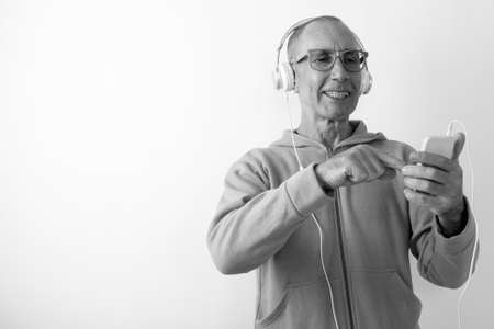 Studio shot of happy bald senior man smiling while using mobile phone and listening to music with eyeglasses against white backgroundの写真素材