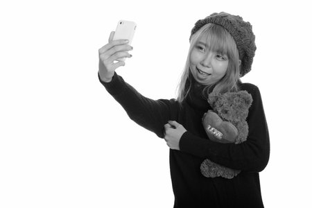 Young happy Asian woman smiling and holding teddy bear with heart and love sign while taking selfie with mobile phoneの写真素材