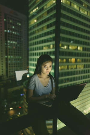 Young beautiful Asian woman using laptop against glass window with view of the city at nightの写真素材