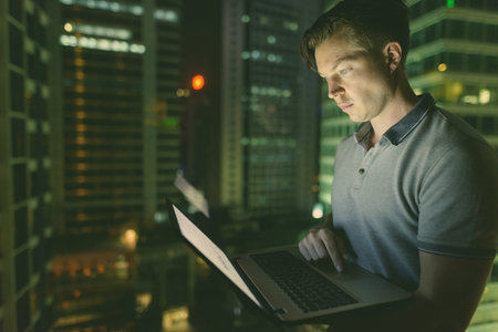 Young handsome man using laptop against glass window with view of the city at night