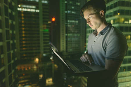 Young handsome man using laptop against glass window with view of the city at nightの写真素材