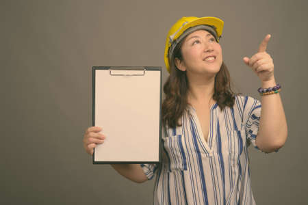 Mature beautiful Asian businesswoman wearing hardhat against gray backgroundの写真素材