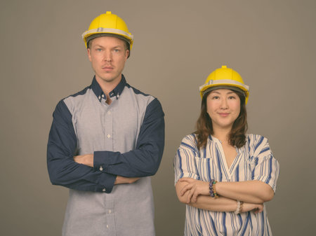 Young handsome businessman and mature Asian businesswoman wearing hardhat against gray background