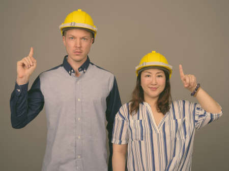 Young handsome businessman and mature Asian businesswoman wearing hardhat against gray backgroundの写真素材
