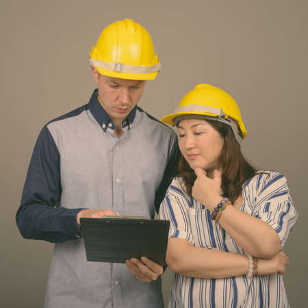 Young handsome businessman and mature Asian businesswoman wearing hardhat against gray backgroundの写真素材
