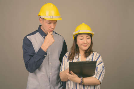 Young handsome businessman and mature Asian businesswoman wearing hardhat against gray backgroundの写真素材