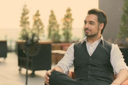 Young handsome Indian man thinking while sitting at rooftop restaurant in Bangkok Thailand