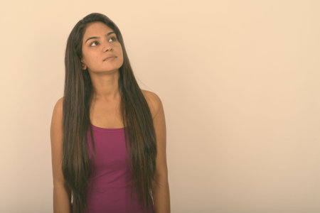 Studio shot of young Indian woman thinking while looking up and wearing sleeveless against white...