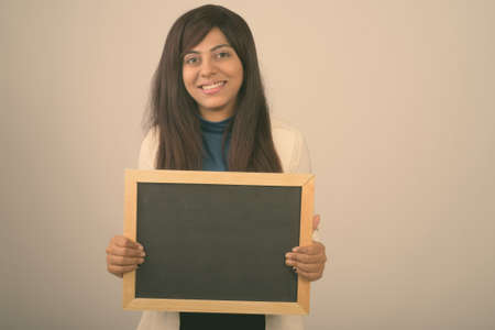Studio shot of young happy Persian businesswoman smiling while holding blank blackboard against gray backgroundの写真素材