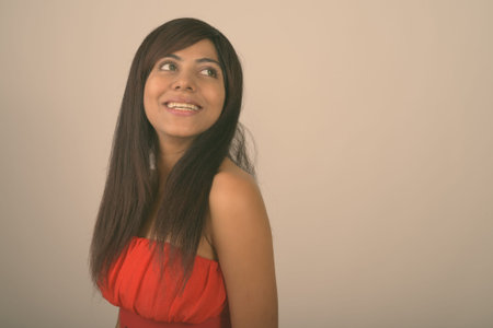 Close up of young happy Persian woman smiling while thinking while looking back in red dress against gray backgroundの写真素材