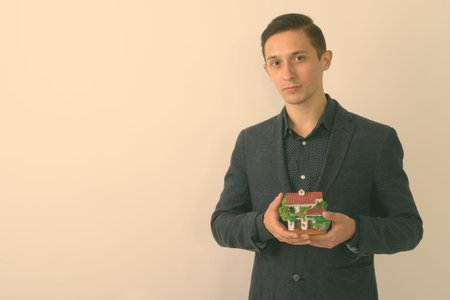 Studio shot of young handsome businessman holding house figurine against white background