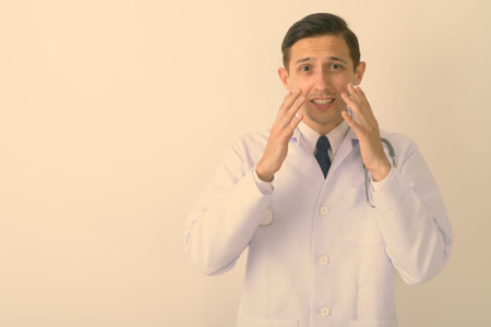 Studio shot of young handsome man doctor looking shocked against white background