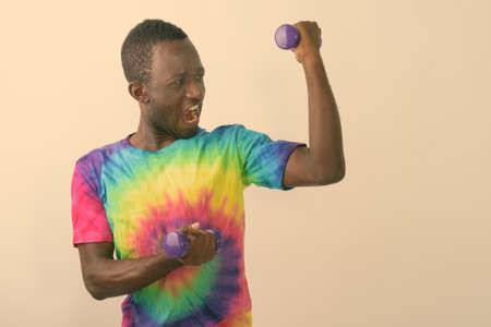 Studio shot of young black African man feeling powerful while holding light weight dumbbells and flexing muscles against white backgroundの写真素材