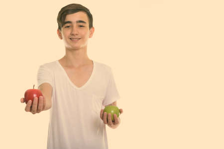 Studio shot of young happy Persian teenage boy smiling while giving red apple and holding green appleの写真素材