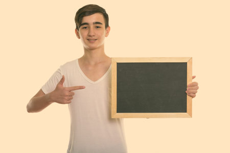 Studio shot of young happy Persian teenage boy smiling while holding and pointing at blank...