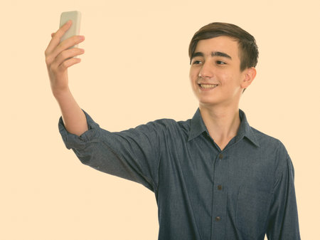 Studio shot of young happy Persian teenage boy smiling while taking selfie picture with mobile phone