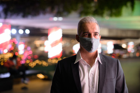 Businessman wearing suit and protective facial mask in city at night while thinking