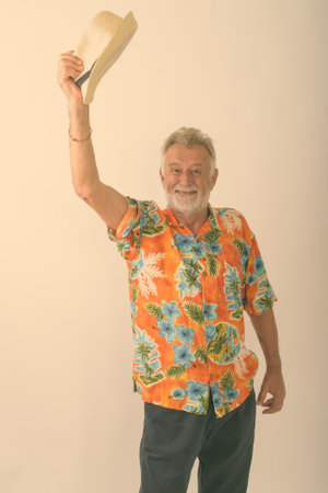 Studio shot of happy senior bearded tourist man smiling and standing while raising hat to greet against white backgroundの写真素材