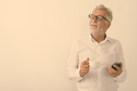 Studio shot of thoughtful happy senior bearded man smiling while holding coffee cup and mobile phone with eyeglasses against white backgroundの写真素材