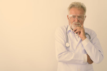 Studio shot of handsome senior bearded man doctor thinking while wearing protective glasses against white backgroundの写真素材