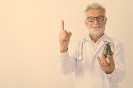 Studio shot of handsome senior bearded man doctor holding avocado while pointing finger up against...