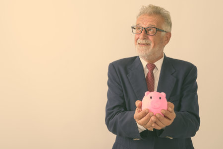 Studio shot of happy senior bearded businessman smiling while thinking and holding piggy bank against white backgroundの写真素材