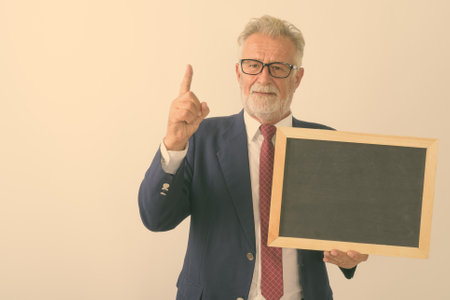 Studio shot of handsome senior bearded businessman pointing finger up while holding blank blackboard against white backgroundの写真素材
