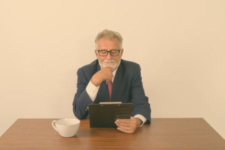 Studio shot of handsome senior bearded businessman thinking while reading on clipboard with coffee cup on wooden table against white backgroundの写真素材