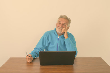 Studio shot of happy senior bearded man smiling and thinking while holding eyeglasses with laptop on wooden table against white backgroundの写真素材