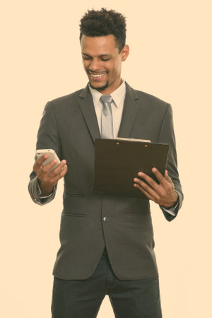 Young happy African businessman smiling while holding clipboard and using mobile phoneの写真素材
