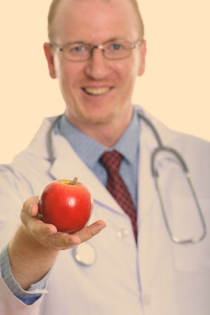 Studio shot of happy man doctor smiling while giving red apple with focus on appleの写真素材