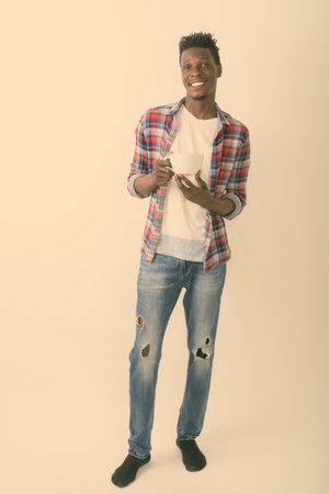 Full body shot of young happy black African man smiling and standing while holding coffee cup against white backgroundの写真素材