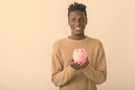 Studio shot of young happy black African man smiling while holding piggy bank against white backgroundの写真素材