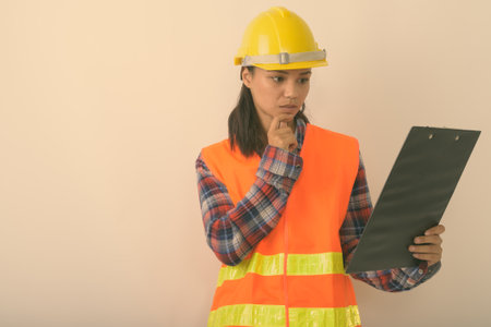 Studio shot of young Asian woman construction worker reading on clipboard while thinking against white backgroundの写真素材