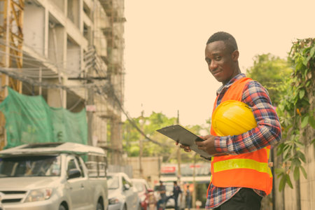 Young black African man construction worker holding clipboard and hard hat at building site